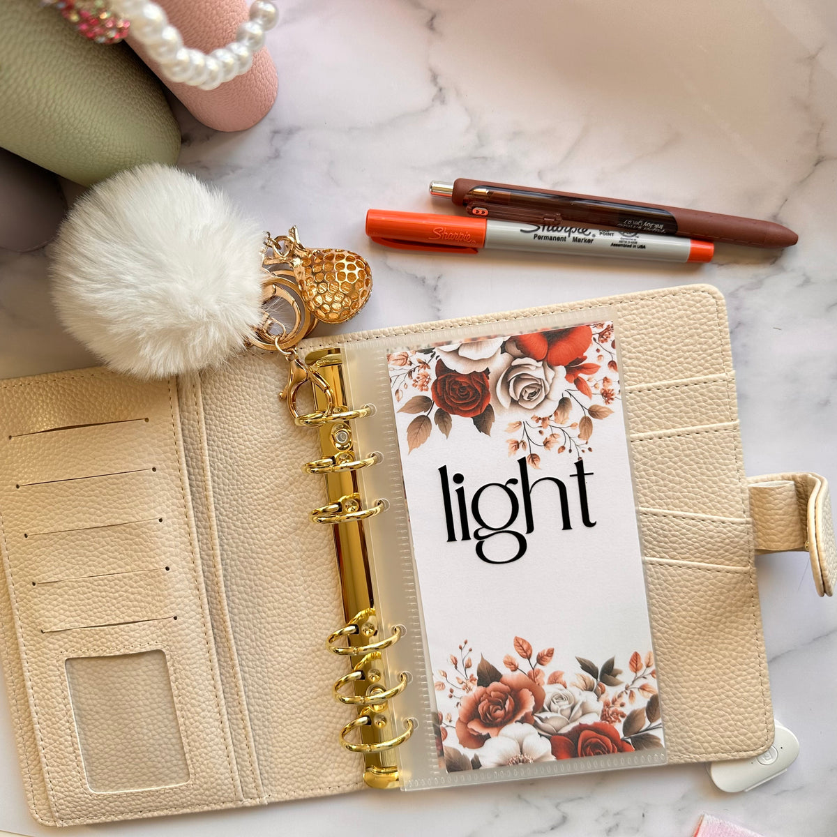 Beige planner with floral design, gold ring, and decorative keychain on a marble surface.